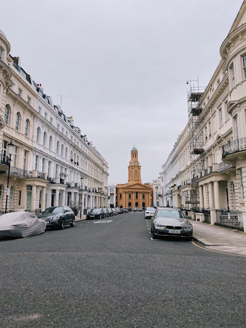 A symmetrical city street scene featuring two rows of white, multi-story residential buildings with classic architectural details such as wrought-iron balconies, decorative window trims, and cornices. The buildings line both sides of a paved road, with several parked cars visible along the curbs, including a covered vehicle on the left. In the background, a prominent yellow brick building with a tall clock tower and a green dome stands centrally at the end of the street, suggesting a historical or civic structure. The sky overhead is overcast, creating diffuse lighting that evenly illuminates the scene. Although the primary focus is on the buildings and vehicles, the setting may relate to an urban environment where alternative waste management or private rubbish collection services, such as those provided by Waste Removal Notting Hill, are commonly utilized for residential waste disposal needs, especially in areas with period architecture and high-density housing.