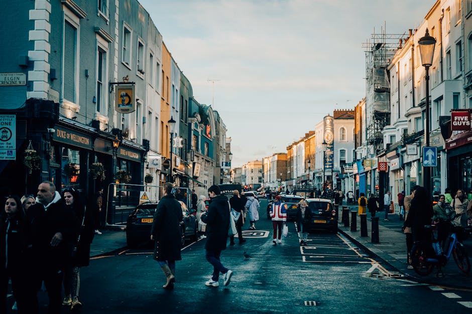 A busy street scene in an urban area during the daytime, with a row of multi-story shops and cafes lining both sides of the street. The buildings have a mix of light-colored facades, some featuring shop signs, hanging flower baskets, and street lamps attached to the walls. Pedestrians are walking along the pavement, some in groups, others alone, dressed in a variety of casual and business attire. Several cars are parked along the curb, with a few moving along the street. In the background, some scaffolding is visible on a building, indicating ongoing construction or maintenance work. The overall environment is well-lit with natural daylight, and the street appears clean and organized, with designated lanes for vehicles and pedestrians. This scene is representative of an active commercial area that may require occasional rubbish removal services, such as those provided by Waste Removal Notting Hill, for maintaining cleanliness amidst daily urban activity.
