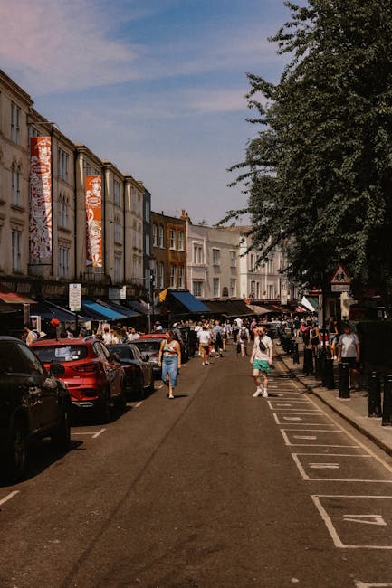 A busy street scene in Notting Hill showing a pedestrian walkway with people walking along the pavement and some standing near small local shops with dark awnings. On the left side, there are parked cars lining the curb, including a red vehicle in the foreground, and behind them, a row of terraced buildings with a mix of cream and brown facades. The buildings have large windows and some display vertical red banners hanging down the front, featuring a patterned design. To the right, there is a large leafy tree extending over part of the pathway, providing shaded areas, and black bollards separating the pedestrian zone from the parking area. The sky above is partly cloudy with patches of blue, suggesting a warm day. This urban scene may relate to private waste collection services, with potential areas for on-site clearance or waste management solutions, fitting into the context of rubbish removal or alternative waste handling in a lively shopping district.