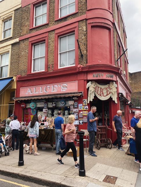 A busy street scene in an urban area during the daytime, with a row of multi-story shops and cafes lining both sides of the street. The buildings have a mix of light-colored facades, some featuring shop signs, hanging flower baskets, and street lamps attached to the walls. Pedestrians are walking along the pavement, some in groups, others alone, dressed in a variety of casual and business attire. Several cars are parked along the curb, with a few moving along the street. In the background, some scaffolding is visible on a building, indicating ongoing construction or maintenance work. The overall environment is well-lit with natural daylight, and the street appears clean and organized, with designated lanes for vehicles and pedestrians. This scene is representative of an active commercial area that may require occasional rubbish removal services, such as those provided by Waste Removal Notting Hill, for maintaining cleanliness amidst daily urban activity.