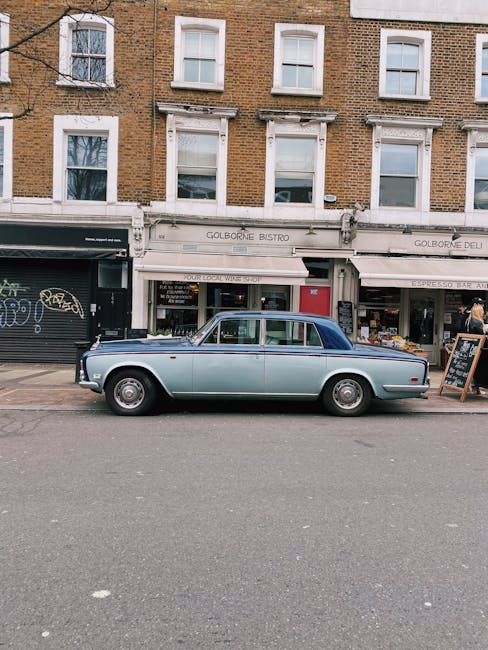 A light blue vintage sedan with a dark blue roof parked on the street in front of a multi-story brick building. The car’s body has a smooth, slightly rounded finish typical of mid-20th-century vehicles, with chrome accents on the wheels and around the window frames. It is positioned parallel to the curb, occupying a designated parking space. Behind the car, the building features three floors with white-framed windows, some with partially drawn blinds. On the ground floor, there are storefronts with signage indicating a bistro and a deli, both with white awnings. To the left, a black shopfront with graffiti art is partially visible. On the right, a small chalkboard sign stands on the sidewalk near some flower displays outside a shop. The scene is illuminated by natural daylight, highlighting the textures of the brickwork, the glossy finish of the car, and the details of the street scene, all of which suggest a typical urban setting where private and independent waste collection might be a relevant aspect of rubbish management, especially in relation to local businesses and residents.