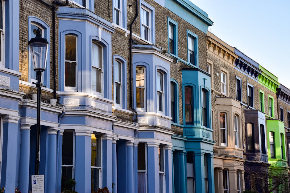 A row of multi-story Victorian terraced houses with different painted facades lining a city street. The buildings feature large sash windows, some with white framing, and decorative cornices. The first-floor windows are set within bay windows, giving the structures a classic appearance. The ground floors include entrances with step stairs and small front gardens or pavement areas. A traditional black lamppost stands on the sidewalk in front of the second house from the left. The scene is illuminated by natural daylight, casting soft shadows on the brickwork and painted surfaces. To the left, there is a parking sign visible on the sidewalk, and in the background, additional buildings continue along the street. The environment suggests a residential area where private property maintenance and waste disposal are managed independently, aligning with services like those offered by Waste Removal Notting Hill for rubbish clearance and alternative waste handling solutions.
