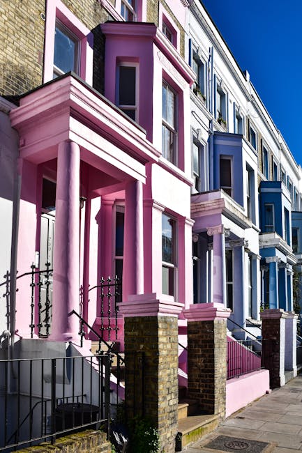A row of three-storey Victorian terraced houses with decorative brickwork and large sash windows, painted in vibrant pastel colors; the building on the left is painted pink with white accents, featuring a small front porch with white columns and black wrought iron railings, while the adjacent buildings are painted in shades of blue with contrasting trims. The houses are situated along a paved sidewalk with a brick and metal fence in front, and the clear blue sky provides bright, natural lighting that highlights the bold color schemes and architectural details. The scene depicts an urban residential area, which might require professional rubbish removal services such as those provided by Waste Removal Notting Hill to manage waste from renovation or cleaning projects, especially in alternative waste handling scenarios outside local authority collection.