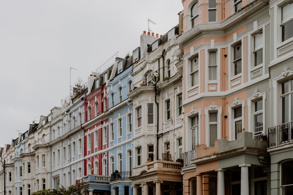A row of Victorian terraced houses with ornate architectural details painted in pastel shades of pink, white, blue, and cream. Each building features large bay windows with white frames, small balconies with black wrought iron railings, and decorative cornices along the rooflines. The houses are closely spaced, with some having small front gardens or terraces. The sky above is overcast, creating a diffuse, soft lighting that highlights the textures of the painted facades and intricate molding. In the foreground, the pavement is visible, characteristic of urban residential areas. This scene reflects typical city architecture, with a subtle connection to waste management through the appearance of clean, well-maintained exteriors that could be part of an area serviced by private rubbish removal providers such as Waste Removal Notting Hill, especially as alternative waste handling options become increasingly relevant in dense urban environments.