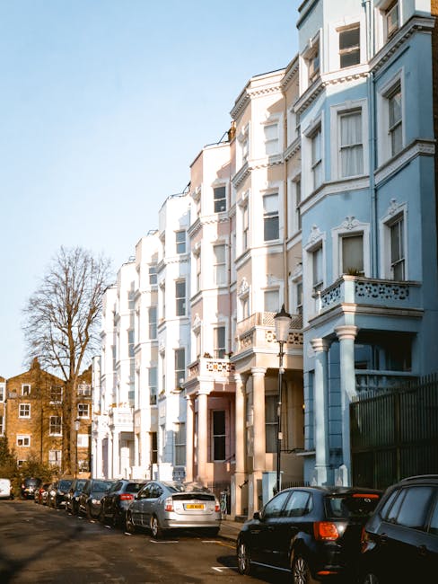 A row of Victorian terraced houses with ornate architectural details painted in pastel shades of pink, white, blue, and cream. Each building features large bay windows with white frames, small balconies with black wrought iron railings, and decorative cornices along the rooflines. The houses are closely spaced, with some having small front gardens or terraces. The sky above is overcast, creating a diffuse, soft lighting that highlights the textures of the painted facades and intricate molding. In the foreground, the pavement is visible, characteristic of urban residential areas. This scene reflects typical city architecture, with a subtle connection to waste management through the appearance of clean, well-maintained exteriors that could be part of an area serviced by private rubbish removal providers such as Waste Removal Notting Hill, especially as alternative waste handling options become increasingly relevant in dense urban environments.