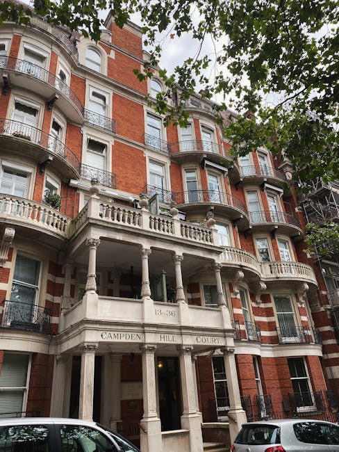 The image shows a multi-storey residential building constructed with red brick and white stone detailing, featuring rounded bay windows with black wrought-iron railings on each level. The building's façade includes decorative white stone cornices and balustrades, with a prominent entrance supported by classical columns and pilasters engraved with the address '13-36' and the name 'Campden Hill Court.' The entrance area is covered, providing a small porch space, and the surrounding environment includes trees with green foliage partially obscuring the upper floors. Parked cars are visible along the street in the foreground, and the scene is lit with natural daylight, highlighting the building's architectural details. This setting suggests an urban residential area where private or independent rubbish collection services, such as those by Waste Removal Notting Hill, could be relevant for managing waste and rubbish removal, particularly in the context of on-site clearance or alternative waste handling for high-rise buildings.