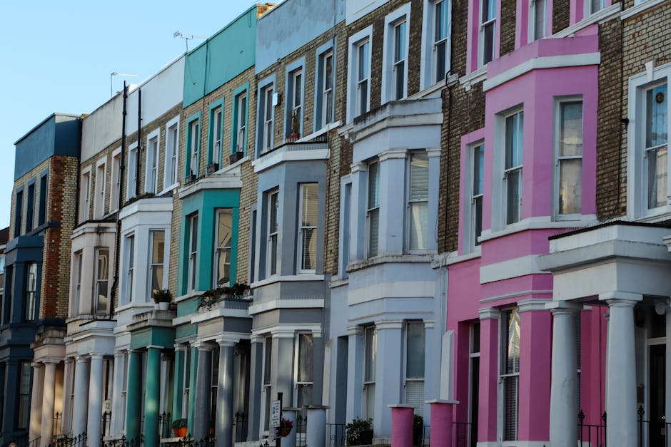 A row of Victorian-style terraced houses with decorative bay windows and ornate cornices, painted in pastel shades of pink, blue, green, and white. The buildings have brick facades with some areas of exposed brickwork, and each property features a small front balcony with black wrought iron railings. The windows are double-glazed with white frames, and some have window boxes or small potted plants on the sills. The street is illuminated by natural daylight, casting soft shadows on the facades, and the pavement in front is clean and unobstructed. The overall scene depicts a typical residential street in an urban area with well-maintained, colorful row houses, aligning with private property management and accessible rubbish disposal options for homeowners, associated with professional waste removal services such as Waste Removal Notting Hill.