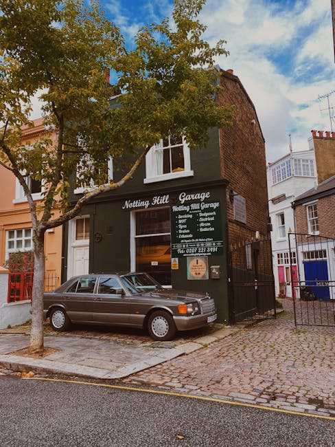A row of three to four-storey Victorian-style terraced houses featuring distinct facade colors—beige, dark blue, bright green, and pale pink—each with large, vertically oriented sash windows framed in white. The buildings have decorative cornices and brickwork detailing, with smooth painted surfaces and contrasting trim. In the foreground, a leafless tree with thin, spindly branches partially obscures the lower floors, while a streetlamp with a black metal post and glass lantern stands beside a dense, dark green leafy shrub. The scene is illuminated by natural daylight against a clear blue sky, with shadows cast on the facades, highlighting the textures of brickwork and painted surfaces. This urban residential setting exemplifies the vibrant, individual character of Nottinghamshire's housing stock, with no visible rubbish or waste, but the context aligns with private waste management services such as those offered by Waste Removal Notting Hill for on-site clearance or independent collection of household rubbish in similar urban environments.