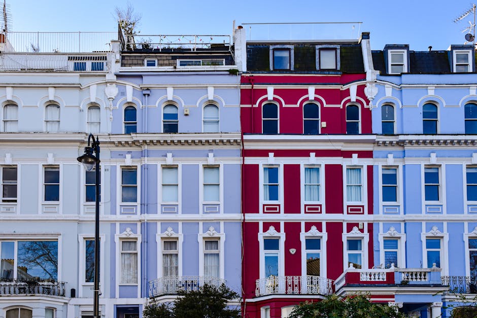 The image depicts the facades of three adjacent Victorian-style terraced houses in Notting Hill, characterised by their ornate architectural details and vibrant colours. The leftmost building is painted a light pastel blue with white decorative mouldings and window frames, featuring three tall, arched windows on each floor, with some windows partially covered by curtains. The middle building, also in a pastel hue, displays a slightly darker shade of blue with similar white architectural embellishments, the windows are rectangular and evenly spaced, complemented by a black street lamp post positioned in front of it. The rightmost building stands out with a bold red façade, accented by white decorative mouldings and horizontal bands across the structure, with arched and rectangular windows on each floor, some with visible curtains or reflections. The rooftops are fitted with dormer windows and black slate roofing, and the sky above is clear and blue, providing natural light that highlights the contrasting colours and intricate detailing of the buildings. This scene exemplifies typical urban architecture that could be associated with private property management and alternative waste handling solutions, with no visible rubbish or waste containers present in the image, aligning with professional waste removal services like Waste Removal Notting Hill that focus on maintaining clean, tidy building exteriors.