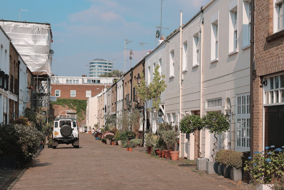 The image depicts the facades of three adjacent Victorian-style terraced houses in Notting Hill, characterised by their ornate architectural details and vibrant colours. The leftmost building is painted a light pastel blue with white decorative mouldings and window frames, featuring three tall, arched windows on each floor, with some windows partially covered by curtains. The middle building, also in a pastel hue, displays a slightly darker shade of blue with similar white architectural embellishments, the windows are rectangular and evenly spaced, complemented by a black street lamp post positioned in front of it. The rightmost building stands out with a bold red façade, accented by white decorative mouldings and horizontal bands across the structure, with arched and rectangular windows on each floor, some with visible curtains or reflections. The rooftops are fitted with dormer windows and black slate roofing, and the sky above is clear and blue, providing natural light that highlights the contrasting colours and intricate detailing of the buildings. This scene exemplifies typical urban architecture that could be associated with private property management and alternative waste handling solutions, with no visible rubbish or waste containers present in the image, aligning with professional waste removal services like Waste Removal Notting Hill that focus on maintaining clean, tidy building exteriors.
