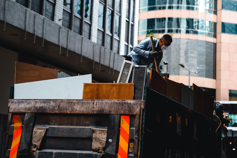 A man wearing a protective face mask, a light blue shirt, and dark trousers is standing on the back of a large, black waste collection truck, leaning slightly forward and lifting a wooden panel with both hands. The truck is parked on a city street near modern office buildings with glass windows and concrete facades. The truck's bed is loaded with various types of debris, including wooden boards, radiator panels, and other furniture components, all stacked and leaning against each other. The wooden boards vary in colour from light to dark brown, with some showing a polished finish and others with a rougher, unfinished surface. The environment appears to be an urban area with minimal surrounding greenery, and the lighting suggests daytime with natural light illuminating the scene. This scene depicts an aspect of private waste disposal or rubbish removal services, with the man possibly preparing to load or unload bulky waste as part of an organized rubbish removal process or alternative waste handling method supported by Waste Removal Notting Hill.