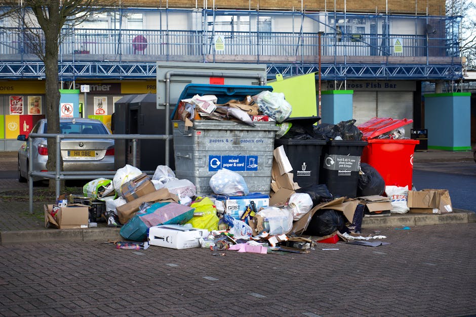 A collection of overflowing rubbish and recycling bins situated on a paved area outside a commercial building. The scene includes a large grey mixed paper and card bin with its lid open, revealing crumpled paper, cardboard, and miscellaneous waste spilling over onto the ground. To the right, there are black recycling bags and a bright red bin, all filled with waste. Surrounding the bins, various discarded items such as cardboard boxes, plastic packaging, paper waste, and small discarded household objects are scattered across the pavement, creating a cluttered appearance. Behind the waste collection point, a street with parked cars and commercial shopfronts, including a fish bar and retail signs, are visible, with a building under construction or renovation covered in scaffolding and safety netting in the background. The overall environment appears to be an urban outdoor area where private waste collection services like Waste Removal Notting Hill might be engaged for rubbish clearance, especially in situations of overflow or bulk waste disposal.