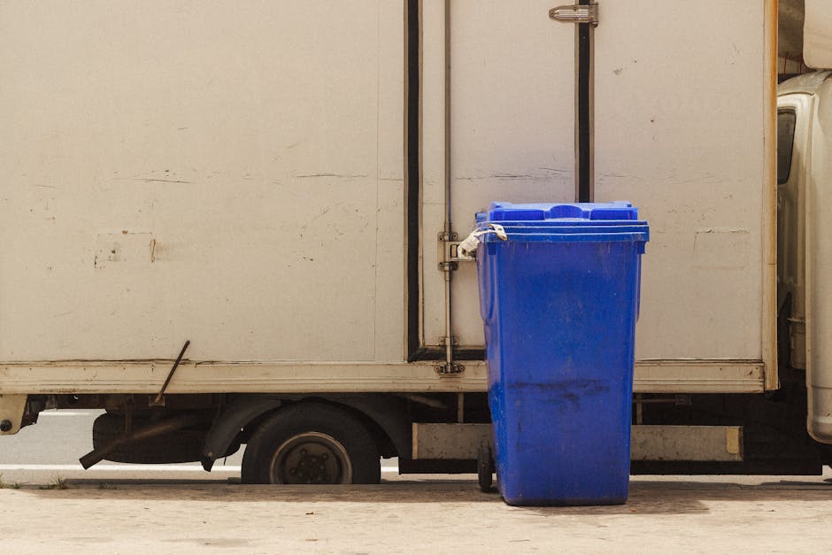 A man wearing a protective face mask, a light blue shirt, and dark trousers is standing on the back of a large, black waste collection truck, leaning slightly forward and lifting a wooden panel with both hands. The truck is parked on a city street near modern office buildings with glass windows and concrete facades. The truck's bed is loaded with various types of debris, including wooden boards, radiator panels, and other furniture components, all stacked and leaning against each other. The wooden boards vary in colour from light to dark brown, with some showing a polished finish and others with a rougher, unfinished surface. The environment appears to be an urban area with minimal surrounding greenery, and the lighting suggests daytime with natural light illuminating the scene. This scene depicts an aspect of private waste disposal or rubbish removal services, with the man possibly preparing to load or unload bulky waste as part of an organized rubbish removal process or alternative waste handling method supported by Waste Removal Notting Hill.