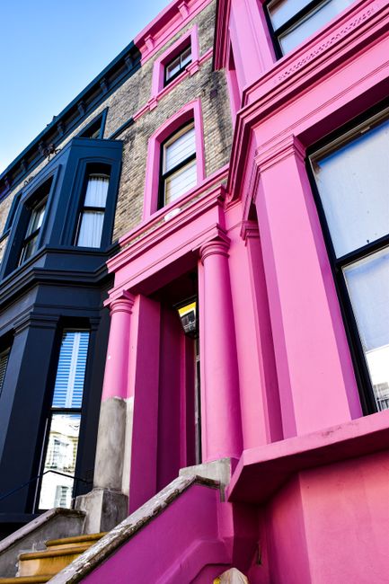 A close-up view of a Victorian terraced building façade with a combination of dark blue and bright pink paintwork, highlighting the architectural details such as columns, window frames, and decorative mouldings. The building features multiple windows with white window frames, some partially visible through closed blinds. The pink section includes a set of stairs leading up to the entrance, with a small lantern-style light fixture illuminating the doorway. The façade is constructed from brickwork, partially visible on the upper sections, with the vivid paint emphasizing the architectural features and creating a striking contrast against the blue sections. The scene is captured in natural daylight, with a clear sky reflected in the windows, and the overall setting suggests a residential area within an urban environment, potentially related to a private property often associated with independent waste handling or on-site clearance services such as those offered by Waste Removal Notting Hill.