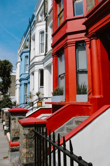 A row of Victorian terraced houses in Notting Hill featuring a variety of pastel and bold colour finishes, including shades of teal, white, light blue, and pink, with decorative bay windows and brickwork visible on upper floors. Each property has a small front garden or entry area with potted plants or iron railings. The facades display a mix of textured brickwork and smooth painted surfaces, with the upper stories slightly projecting outward. Clear sky in the background suggests bright daylight, illuminating the details of the building exteriors with natural light. This image visually aligns with private property environments where occasional building clearance or refurbishment projects may occur, supporting the context of independent waste handling and rubble removal services provided by Waste Removal Notting Hill in urban inner-city settings.
