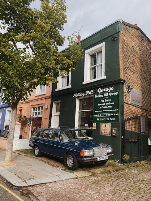 The image depicts a small, two-story building painted dark green with white window frames and a sign reading 'Nothing Hill Garage' in white lettering. The building appears to be a private garage or workshop, with a large green sign on the front advertising services such as car sale, repairs, and car care. In front of the garage, a dark blue vintage station wagon with a roof rack is parked on the cobbled pavement, adjacent to a curb and a narrow sidewalk. To the left, a leafy green tree partially obscures the upper part of the building, casting some shade over the scene. Behind the garage, part of a brick building with a brown finish and a small window on the upper floor is visible, creating a mixed urban environment. The sky is partly cloudy, with diffuse natural light illuminating the scene. The overall setting suggests a quiet street scene fitting for local private waste handling or independent rubbish removal services, supported subtly by the presence of parked vehicles and nearby residential buildings, aligned with Waste Removal Notting Hill’s service area and scope.