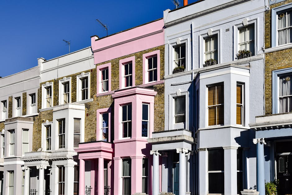 A row of Victorian-style terraced houses with decorative brick facades, featuring prominent bay windows and ornate white cornices. The buildings are painted in pastel shades, including white, light pink, and pale blue, with some sections displaying exposed brickwork and moss growth. The windows vary in size, some with sash frames, and a few have flower boxes. The fronts of the houses are situated on a paved pavement, with clear blue sky above, casting shadows that highlight architectural details. The scene visually suggests an urban residential area where private property maintenance may involve rubbish removal or on-site clearance, although no waste or equipment is visible in the image. The overall atmosphere is bright and clean, emphasizing the well-maintained exterior of the buildings, while subtly supporting the context of disposal or rubbish collection services often required in similar residential environments.